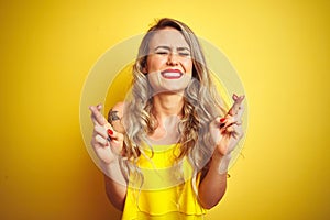Young attactive woman wearing t-shirt standing over yellow  background gesturing finger crossed smiling with hope and eyes