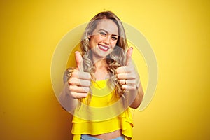 Young attactive woman wearing t-shirt standing over yellow isolated background approving doing positive gesture with hand, thumbs