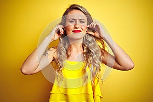 Young attactive woman wearing t-shirt standing over yellow isolated background covering ears with fingers with annoyed expression