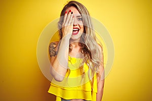 Young attactive woman wearing t-shirt standing over yellow isolated background covering one eye with hand, confident smile on face