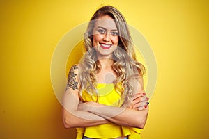 Young attactive woman wearing t-shirt standing over yellow isolated background happy face smiling with crossed arms looking at the