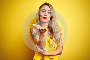 Young attactive woman wearing t-shirt standing over yellow isolated background looking at the camera blowing a kiss with hand on