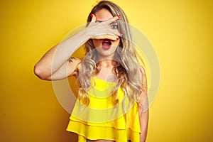 Young attactive woman wearing t-shirt standing over yellow isolated background peeking in shock covering face and eyes with hand,