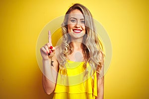 Young attactive woman wearing t-shirt standing over yellow isolated background showing and pointing up with finger number one