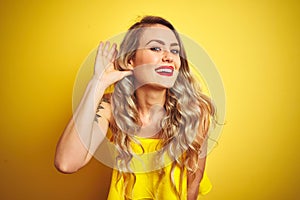 Young attactive woman wearing t-shirt standing over yellow isolated background smiling with hand over ear listening an hearing to