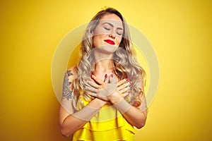 Young attactive woman wearing t-shirt standing over yellow isolated background smiling with hands on chest with closed eyes and