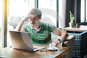 Young attentive businessman in green t-shirt sitting and working on laptop, holding hand on his forehead and looking to something