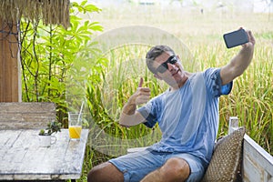 Young attractive 30s Caucasian man smiling happy and relaxed sitting at rice field coffee shop in Asia trip taking selfie picture