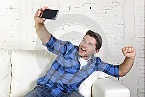 Young attractive 30s man taking selfie picture or self video with mobile phone at home sitting on couch smiling happy