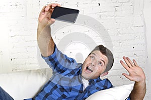 Young attractive 30s man taking selfie picture or self video with mobile phone at home sitting on couch smiling happy