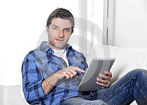 Young attractive 30s man using digital tablet pad lying on couch at home living room networking looking relaxed and happy