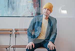 Young attractive adult bearded man hipster in yellow hat sitting on chair and looking at camera, real people lifestyle