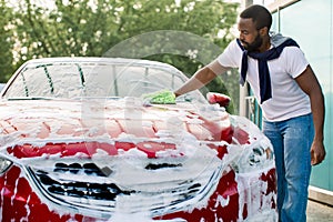 Young attractive African American man washing his red luxury car in a self-service car wash station outdoors with