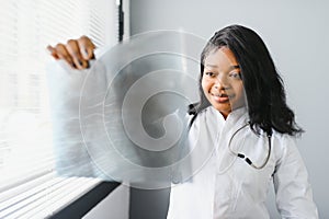 Young attractive african female doctor radiologist looking at x-ray image, while standing in light modern office in