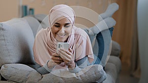 Young attractive arabian muslim girl islamic woman lying on home sofa happy lebanese lady wear hijab relaxing typing