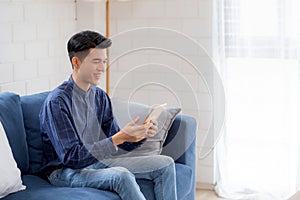 Young attractive asian man resting using browsing tablet computer on sofa at home happy male sitting on couch relax reading