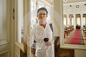 Young attractive asian student in eyeglasses with coffee to go opening library door in university