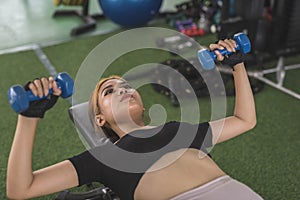 A young and attractive asian woman does bench presses with a pair of blue rubberized dumbbells. Working out chest and upper body