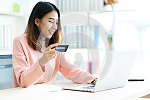 Young attractive asian woman holding credit card sitting at table typing keyboard on laptop computer to shopping online