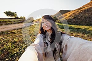 A young attractive Asian woman in a linen dress takes a selfie and laughs outdoors. Smiling Korean woman having fun and taking photo