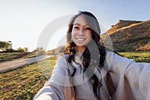 A young attractive Asian woman in a linen dress takes a selfie and laughs outdoors photo