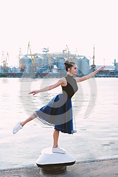 Young attractive ballerina in blue dress on sea coast on background port. Fine art ballet concept. Slim girl dance on sea beach.