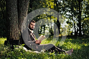 Young attractive bearded business man sitting on green grass under tree and resting in park. Read book, drink coffee