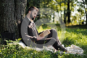 Young attractive bearded business man sitting on green grass under tree and resting in park. Read book, drink coffee