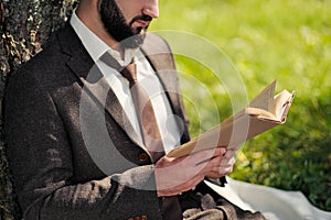 Young attractive bearded business man sitting on green grass under tree and resting in park. Read book, drink coffee