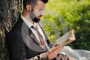 Young attractive bearded business man sitting on green grass under tree and resting in park. Read book, drink coffee