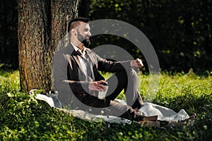 Young attractive bearded business man sitting on green grass under tree and resting in park. Read book, drink coffee