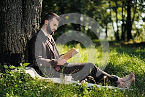 Young attractive bearded business man sitting on green grass under tree and resting in park. Read book, drink coffee