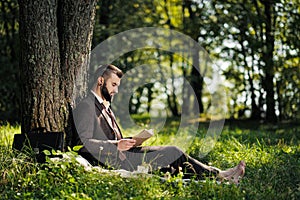 Young attractive bearded business man sitting on green grass under tree and resting in park. Read book, drink coffee