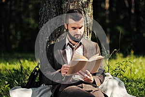 Young attractive bearded business man sitting on green grass under tree and resting in park. Read book, drink coffee