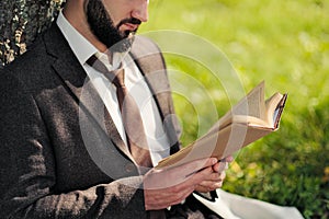 Young attractive bearded business man sitting on green grass under tree and resting in park. Read book, drink coffee