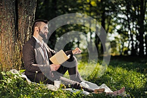 Young attractive bearded business man sitting on green grass under tree and resting in park. Read book, drink coffee