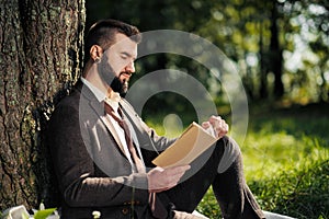 Young attractive bearded business man sitting on green grass under tree and resting in park. Read book, drink coffee