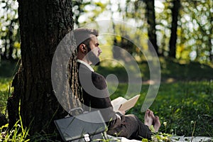 Young attractive bearded business man sitting on green grass under tree and resting in park. Read book, drink coffee