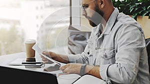 Young attractive bearded hipster man sits in cafe in front of aptop, reading paper documents. Freelancer works remotely