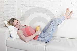 Young attractive blond hair woman holding cup of coffee lying on sofa couch at home living room sleeping relaxed