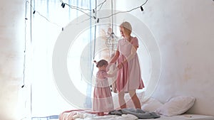 Young attractive blond mom jumping on bed with her little charming daughter in pink dresses.