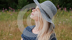 A young attractive blond woman in a blue dress and hat is sitting on a plaid on the grass. The wind blows hair.