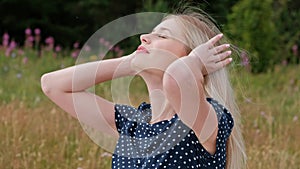 A young attractive blond woman in a blue dress and hat is sitting on a plaid on the grass. The wind blows hair.