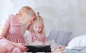 Young attractive blond woman with her little charming daughter in pink dresses seriosly watching something in tablet.