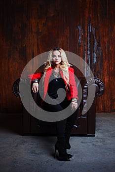 Young and attractive blond woman in red jacket sits in leather armchair, background grunge rusty wall