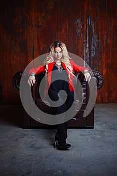 Young and attractive blond woman in red jacket sits in leather armchair, background grunge rusty wall