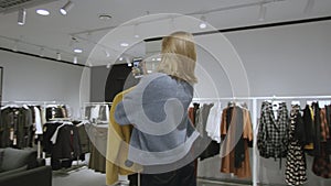 Young attractive blond woman is taking a selfie with a yellow jacket at clothing store, tracking shot, 360 degree