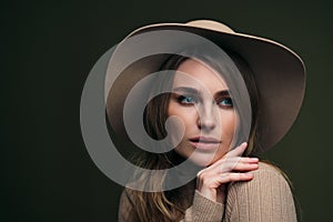 A young attractive blonde woman in a beige sweater, fedora hat and brown leather trousers poses on a dark green olive