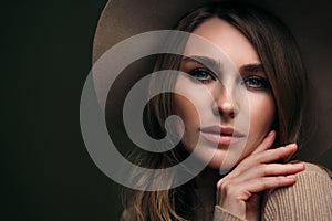 A young attractive blonde woman in a beige sweater, fedora hat and brown leather trousers poses on a dark green olive