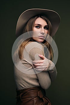 A young attractive blonde woman in a beige sweater, fedora hat and brown leather trousers poses on a dark green olive